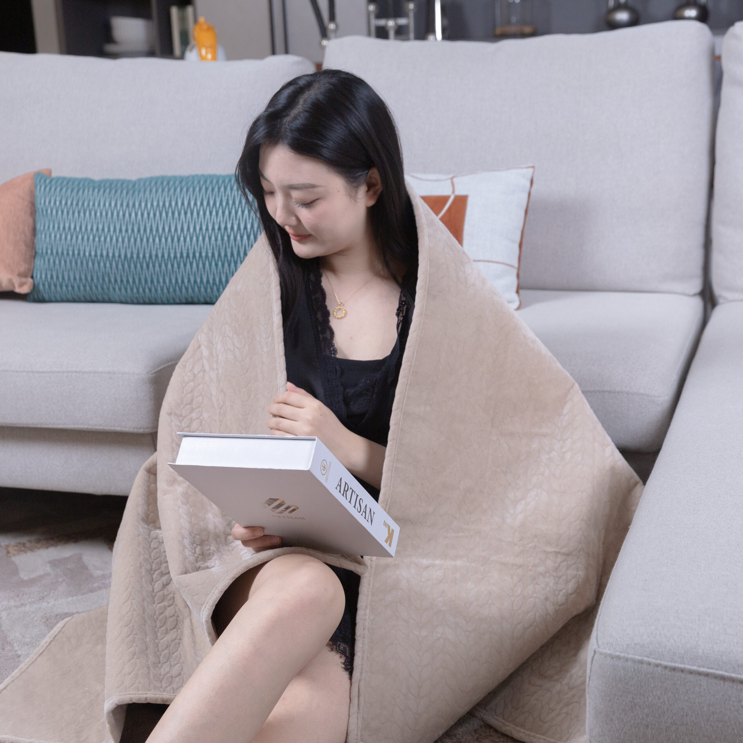 Woman comfortably reading a book wrapped in a cozy blanket on a couch which provide even far infrared heat distribution.